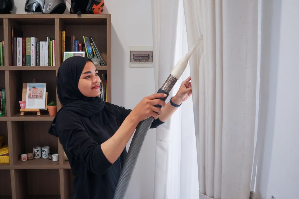 Petugas kebersihan wanita dari King House Cleaning sedang membersihkan gordyn dengan vakum, memastikan kebersihan dan kerapian tirai