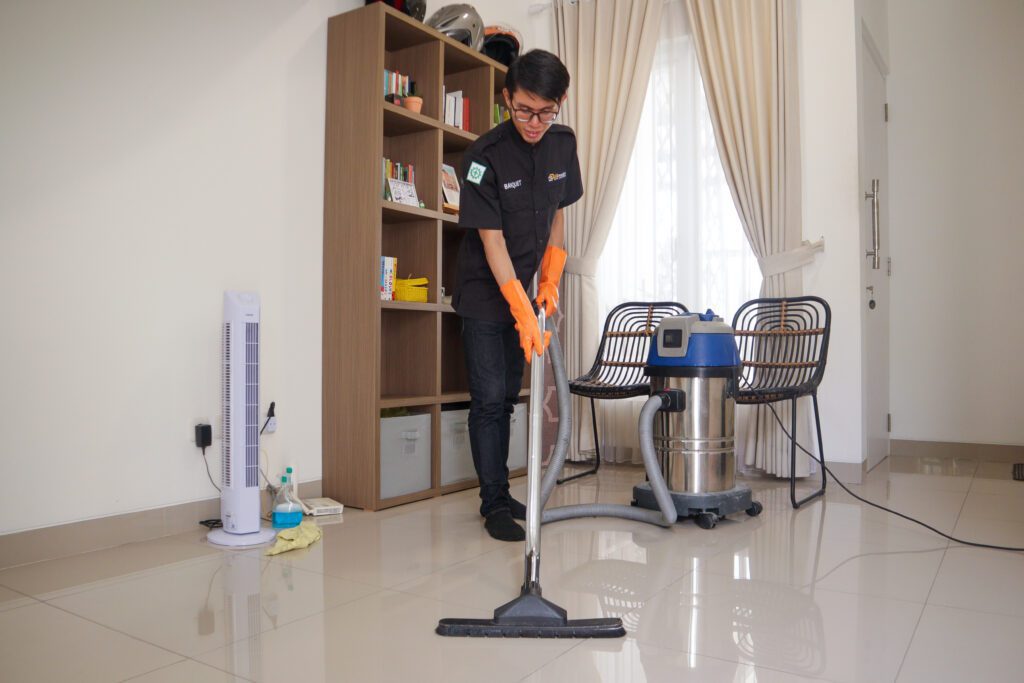 Petugas kebersihan King House Cleaning sedang menggunakan vakum untuk membersihkan lantai, memberikan layanan pembersihan profesional di dalam ruangan.
